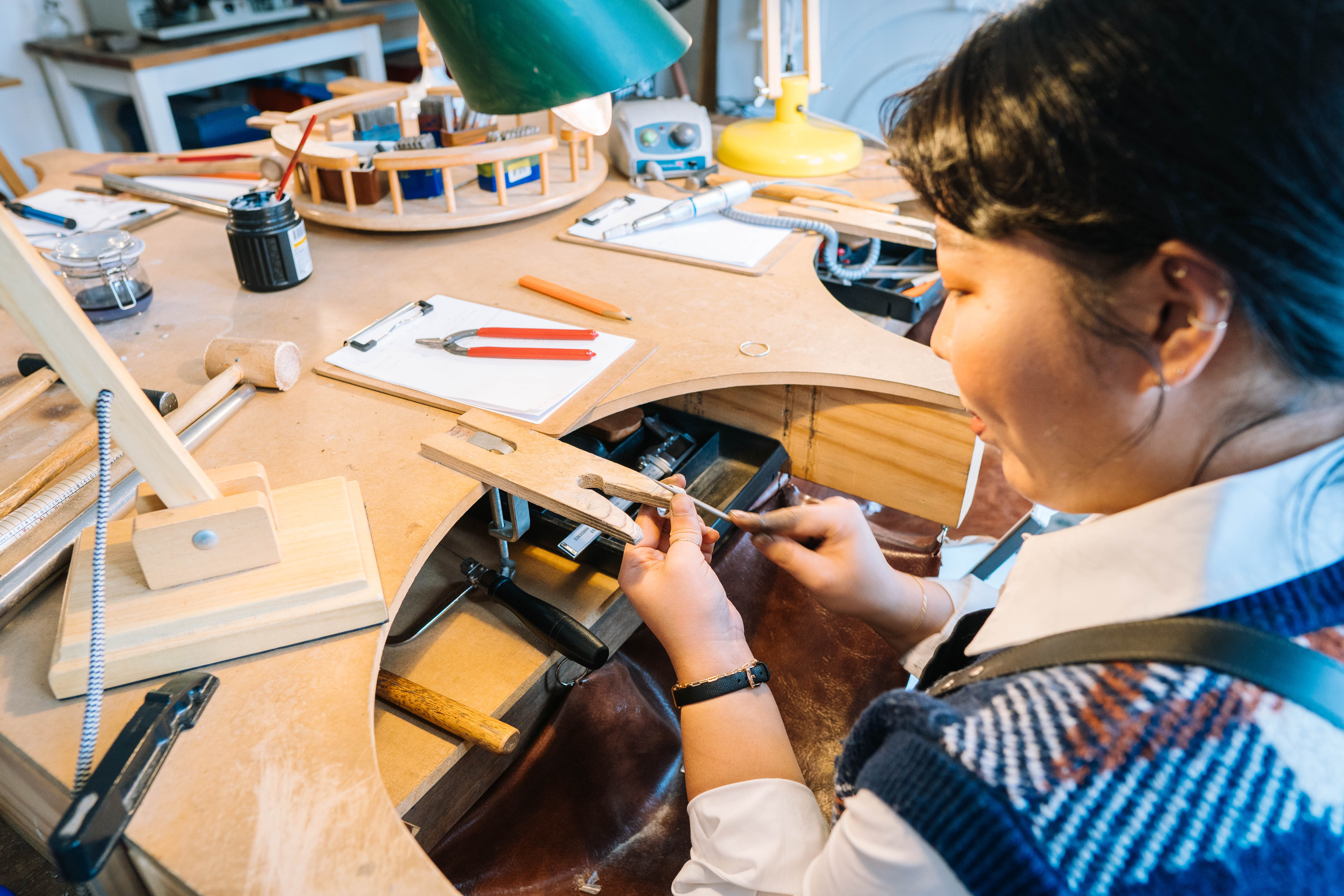 A woman sits at a workbench, using jeweller's tools to make a ring